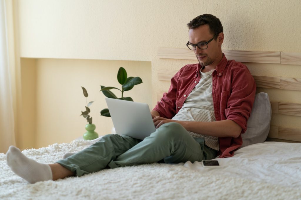 Concentrated Caucasian man sits on bed in hotel room reading news via internet or checking email